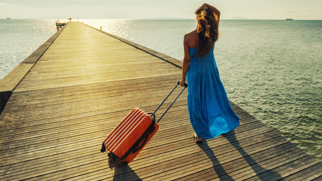 青いロングワンピースを着た女性がスーツケースを引き、海を眺めている