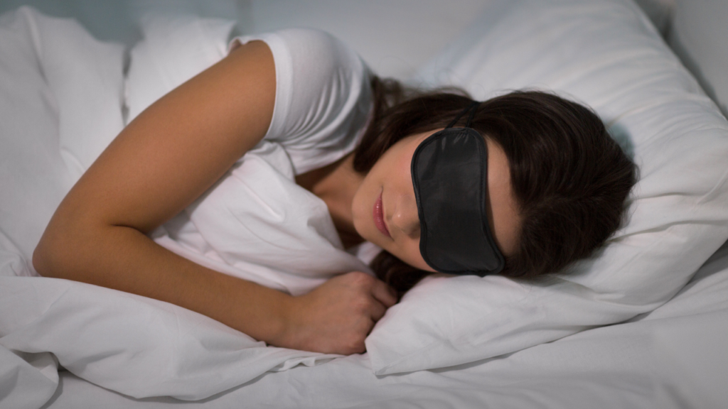 白いベッドで横向きで寝る女性。黒いアイマスクを着用している